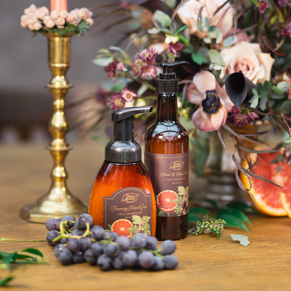 Two bottles of soap on a wooden surface with a candle and flowers in the background