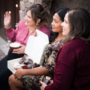 Three women sitting together, enjoying a warm beverage by a stone fireplace.